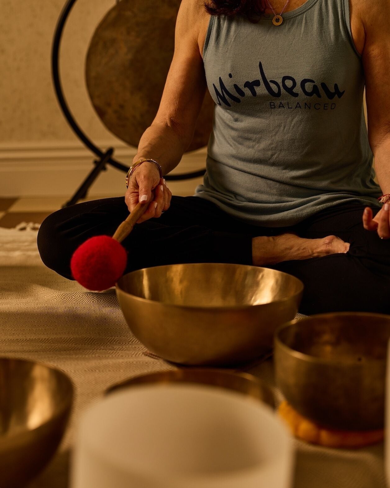 A person sitting on a mat with a large brass bowl and a red mallet, surrounded by other bowls.