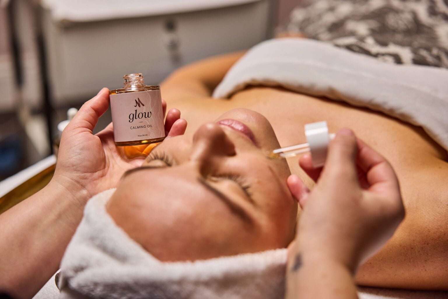 A person receiving a facial treatment with a bottle labeled "glow CALMING OIL".