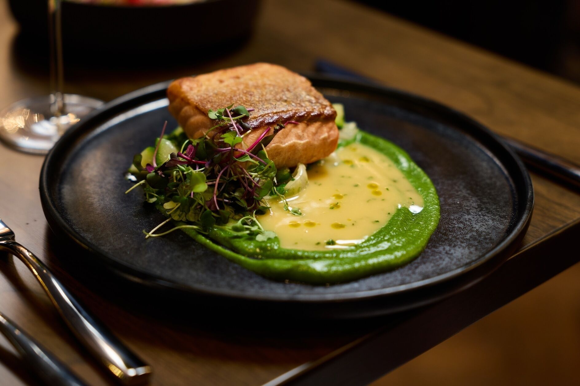 A plate with a piece of salmon, greens, and a sauce on a black plate.