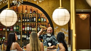 Un bar con un barista che prepara un cocktail, scaffali pieni di liquori e clienti seduti ai tavoli.