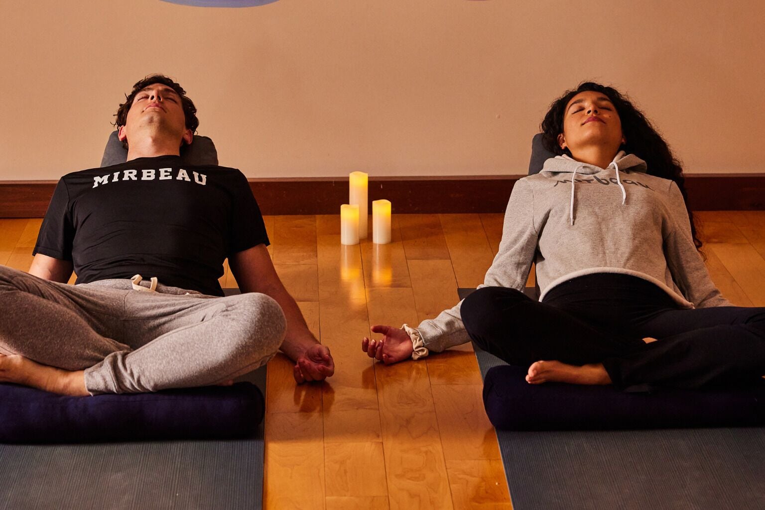 Two people meditating on yoga mats in a room with wooden flooring and candles.