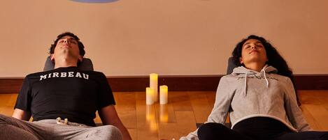 Two people meditating on yoga mats in a room with wooden flooring and candles.