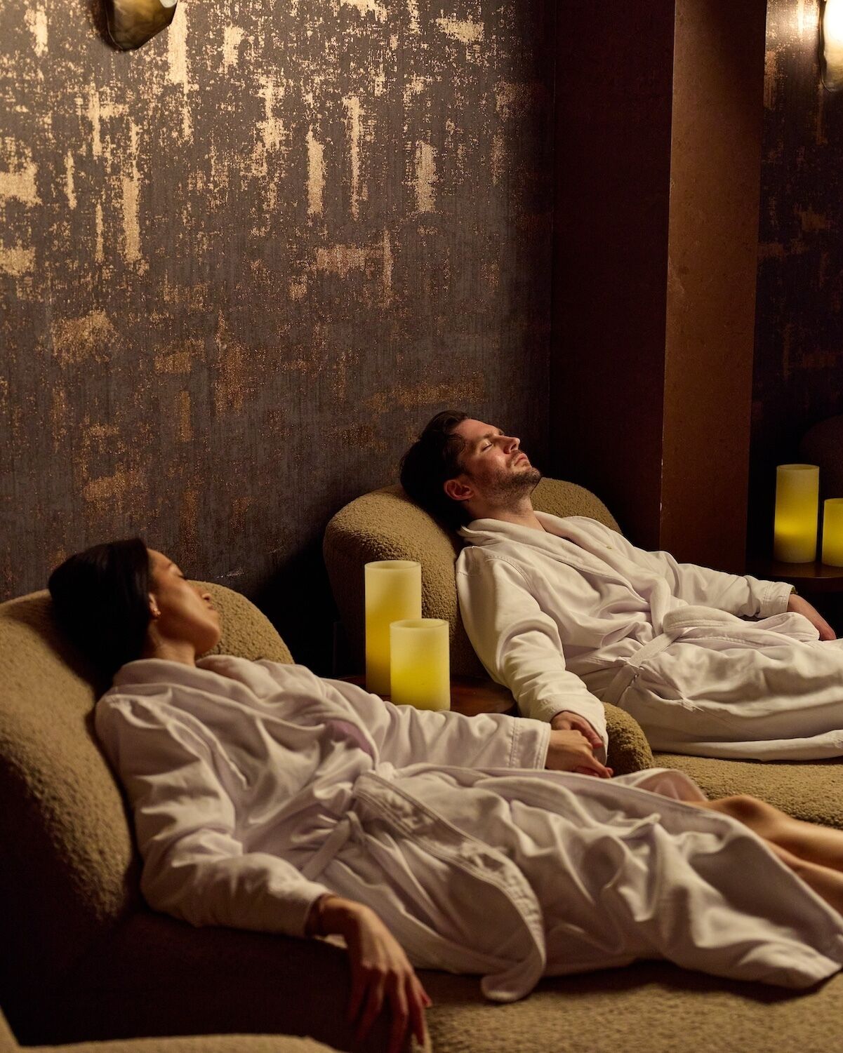 Twee personen liggen op een bed in een kamer met een gestructureerde muur en een zuil.