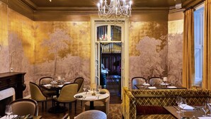 Una sala da pranzo con un lampadario, tavoli apparecchiati e vista su una stanza con parete decorata.