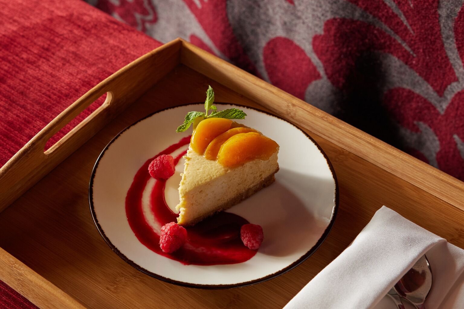 Een stuk cheesecake op een wit bord met rode saus en frambozen, geplaatst op een houten dienblad.