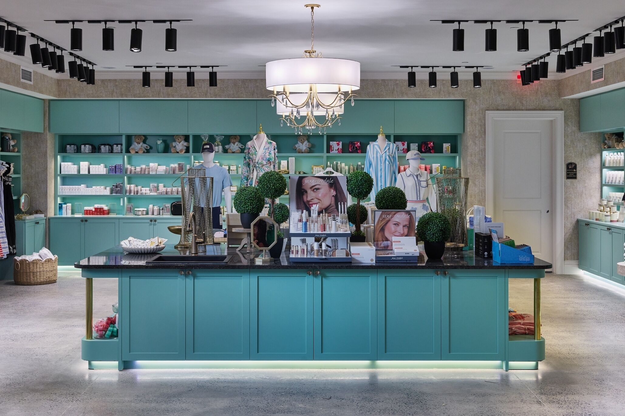 Un hall d’hôtel de charme, avec des meubles de couleur turquoise, un îlot central surmonté d’un lustre, et une variété de produits exposés.