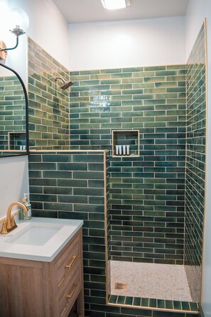Un bagno con doccia rivestita in piastrelle verdi, lavandino bianco con accessori dorati e mobile bagno in legno.