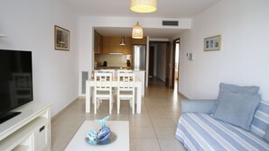 Una sala de estar moderna con una televisión de pantalla plana, un sofá, una mesa de comedor con sillas y una cocina al fondo.