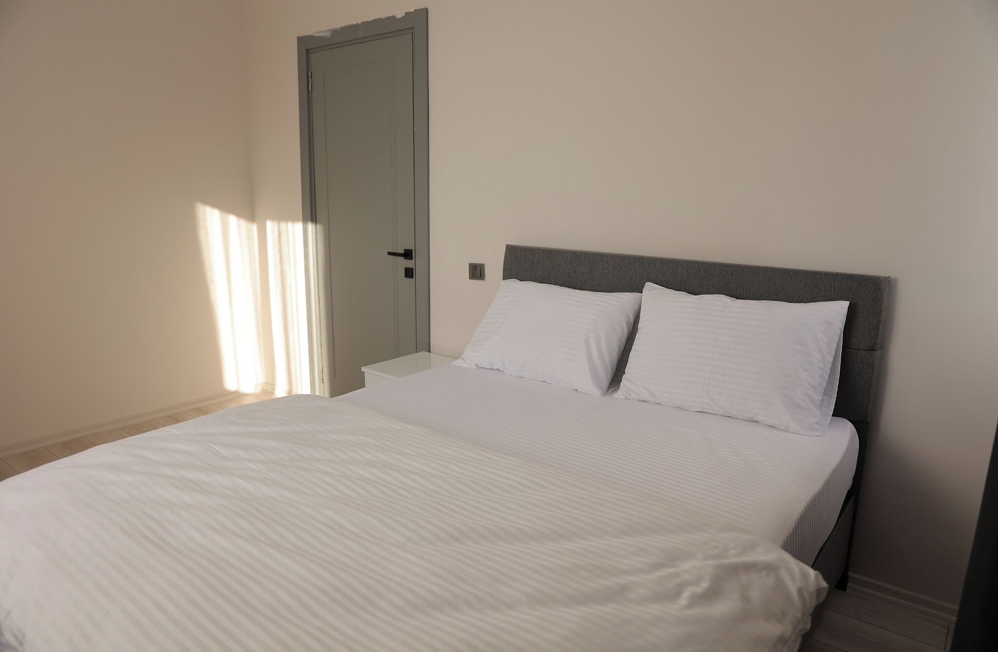 Een slaapkamer met een bed, witte beddengoed, een grijs hoofdeinde, een witte nachtkastje en een deur.
