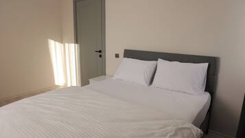 Un dormitorio con una cama, ropa de cama blanca, un cabecero gris, una mesita de noche blanca y una puerta.