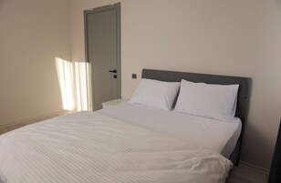 Un dormitorio con una cama, ropa de cama blanca, un cabecero gris, una mesita de noche blanca y una puerta.