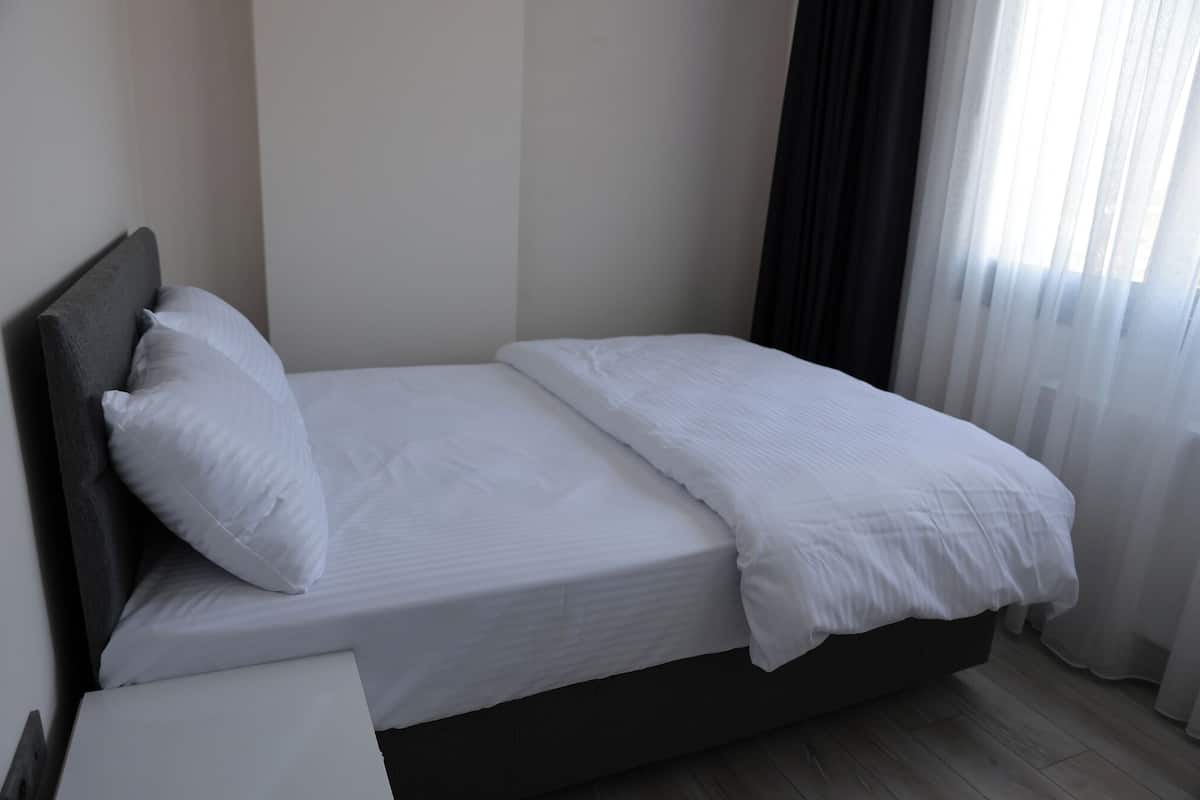 Una cama individual con ropa de cama blanca en una habitación con suelo de madera y una ventana con cortinas de gasa.