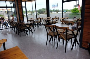Interior de restaurante com várias mesas e cadeiras, amplas janelas e vista para a rua.