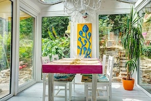 Un invernadero con una mesa de comedor dispuesta para dos personas, rodeada de sillas blancas con cojines de colores, un cuadro abstracto amarillo y una planta en maceta.