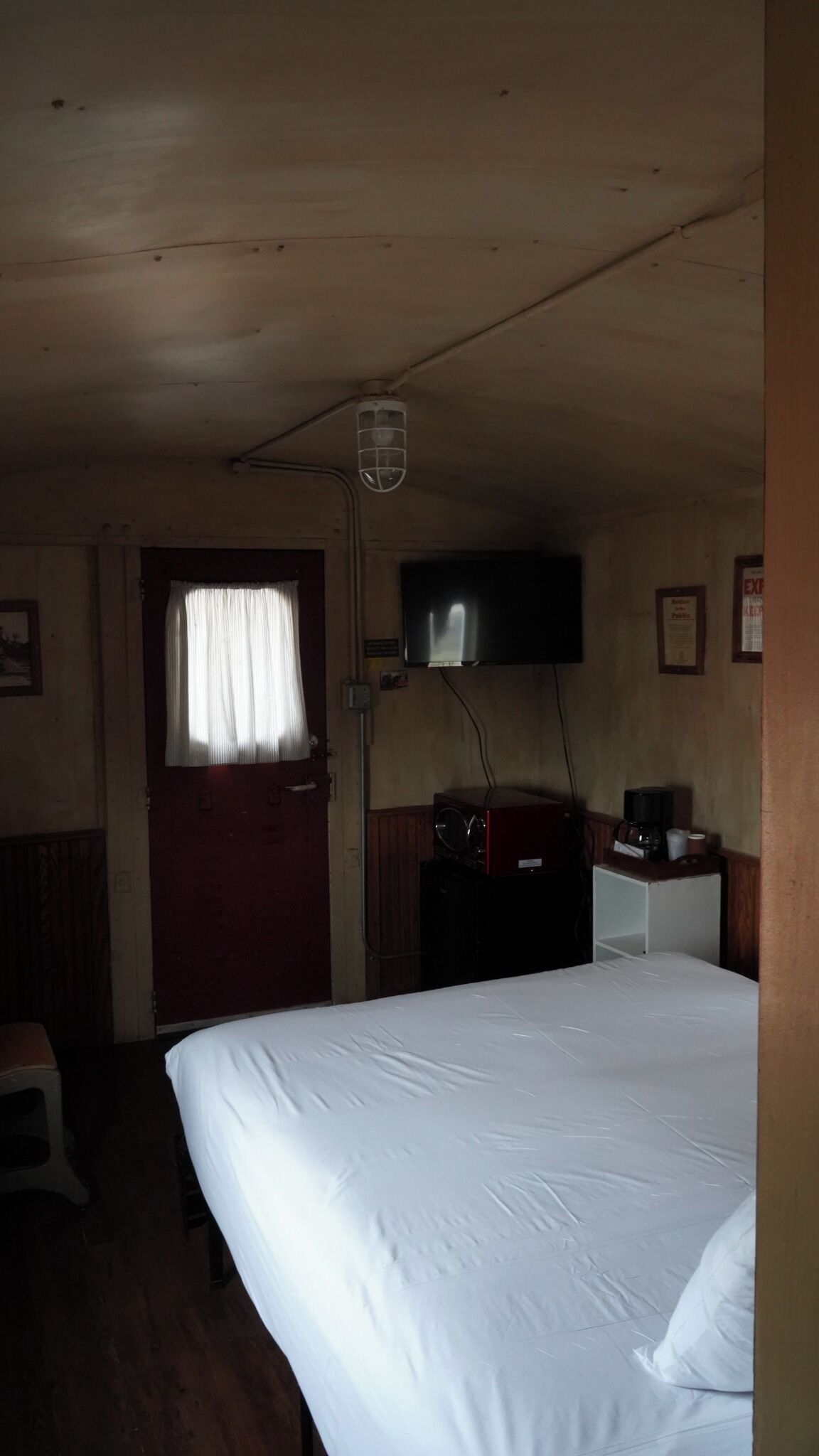 Una habitación pequeña, con poca luz, que tiene una cama individual, un televisor montado en la pared y una puerta de madera con un ventanal.