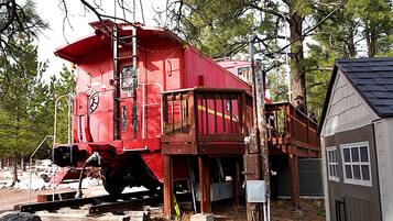 A red train car with a wooden platform and a ladder, surrounded by trees and a small building.