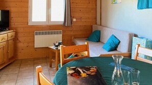 Una cabaña acogedora con paredes de madera, un comedor con una mesa preparada para dos, un sofá con cojines azules y un televisor sobre un mueble de madera.
