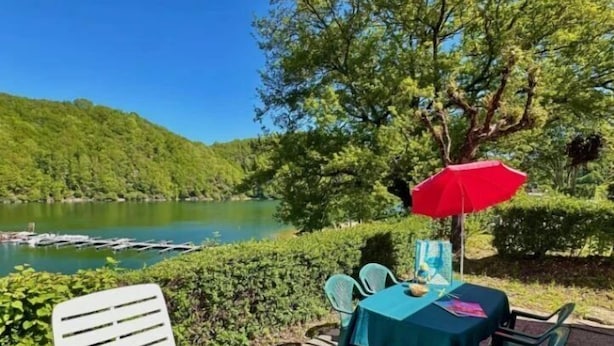 Ein Sitzbereich im Freien mit Tisch und Stühlen, einem roten Sonnenschirm und Blick auf einen See und bewaldete Hügel.