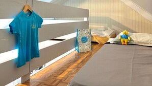 Ein Schlafzimmer mit schrägem Dach, einem Bett mit grauer Matratze, einem hängenden blauen Hemd, einem kleinen Spielente und einer Tasche mit buntem Muster.