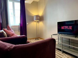 Een woonkamer met een bank, een vloerlamp, een televisie waarop 'NETFLIX' wordt weergegeven, en een raam met gordijnen.