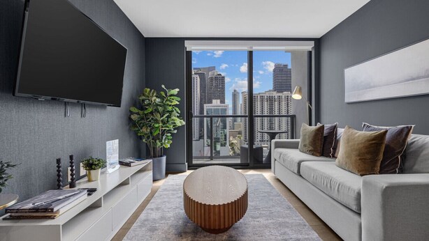 Ein modernes Wohnzimmer mit großem Fernseher, einem grauen Sofa, einem runden Couchtisch und Blick auf die Stadt durch große Fenster.