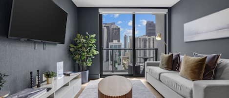Una sala moderna con un televisor grande, un sofá gris, una mesa de centro redonda y vistas a la ciudad a través de amplias ventanas.