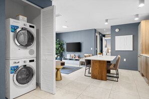 Ett modernt tvättrum med tvättmaskiner och torktumlare staplade på varandra, en matplats med ett träbord och stolar samt en platt-TV som är monterad på väggen.