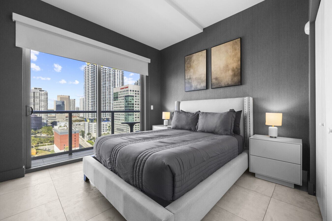 A modern bedroom with a large bed, a nightstand, a lamp, and a view of a cityscape through a sliding door.