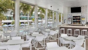 A spacious outdoor seating area with white tables and chairs, a bar with a stone wall and a flat-screen TV, and large windows offering views of palm trees and a pool. - Luxurious 2 BRD/2BATH  Suite in serene Marco Island (Marco Island)