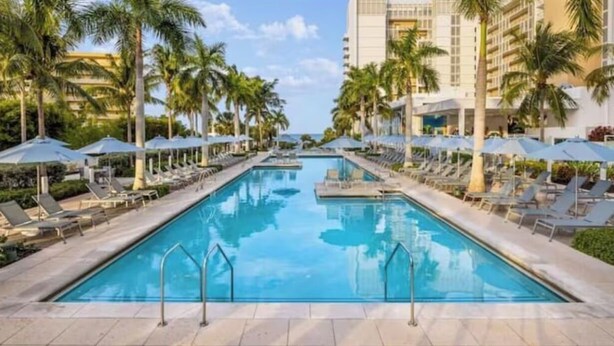 A swimming pool with lounge chairs and umbrellas, surrounded by palm trees and a hotel building. - Luxurious 2 BRD/2BATH  Suite in serene Marco Island (Marco Island)