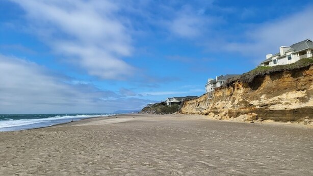 Una playa con casas en un acantilado, cielo despejado y el océano al fondo.