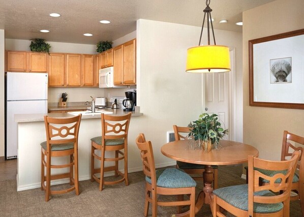 A compact kitchen with wooden cabinets, a white refrigerator, a microwave, and a coffee maker. There is a dining table with four chairs and a yellow pendant light.