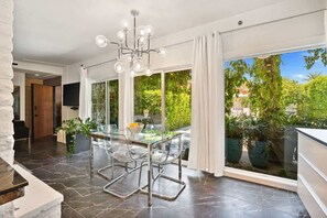 A modern dining area with a glass table and chairs, a chandelier, and a view of greenery outside. - Exclusive use of the property (All 5 units) (Palm Springs)