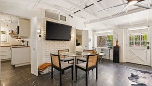 A modern kitchen with white cabinetry, a flat-screen TV mounted on the wall, and a dining area with a round table and chairs. - Adobe Cottage; Unit A (Palm Springs)