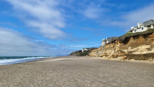 Una playa con casas en un acantilado, cielo despejado y el océano al fondo.