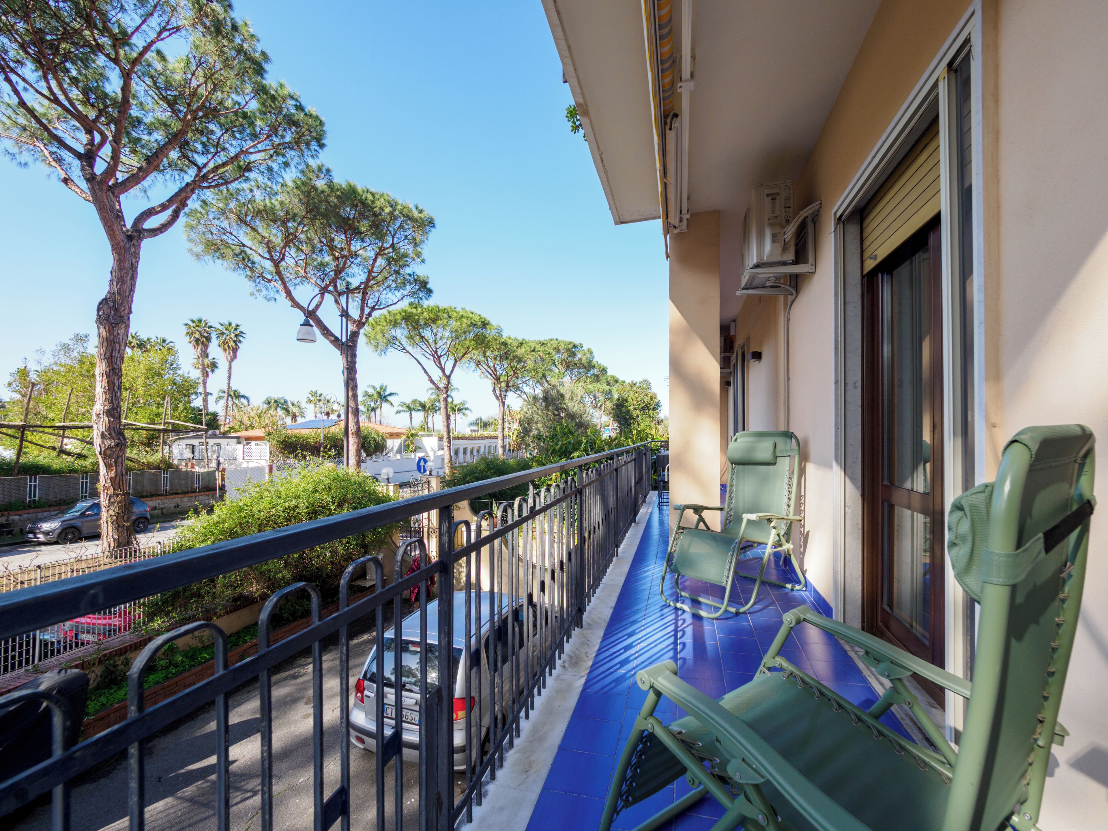 A balcony with a blue floor, two green chairs, a metal railing, and a view of parked cars and trees.