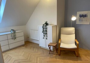 Ein Zimmer mit einem Holzboden im Fischgrätmuster, einem weißen Heizkörper, einem weißen Schrank mit einer Pflanze, einem Holzschemel und einem weißen Stuhl.