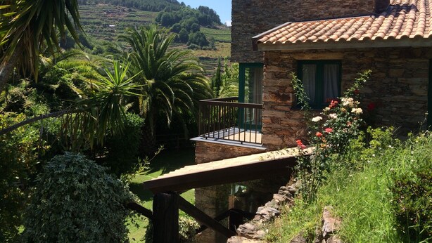 Una casa de piedra con balcón, rodeada de exuberante vegetación y vistas a campos en terrazas.