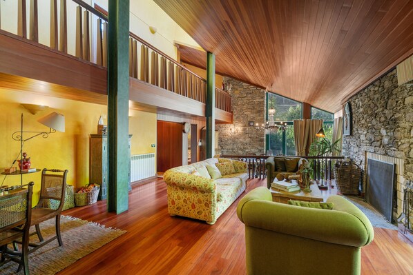 A spacious living area with a high ceiling, wooden beams, a stone wall, and a large window providing a view of the outdoors.
