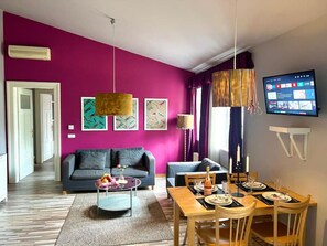 A modern living room with a vibrant pink wall, a grey sofa, a wooden dining table set for four, and a flat-screen TV mounted on the wall. - Prague Dragonfly Apartment (Prague 1)