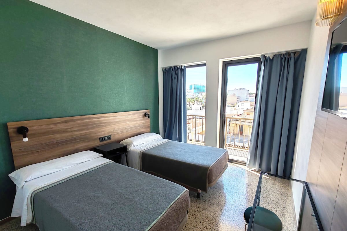 A hotel room with two single beds, a wooden headboard, a small table, and a balcony with curtains.
