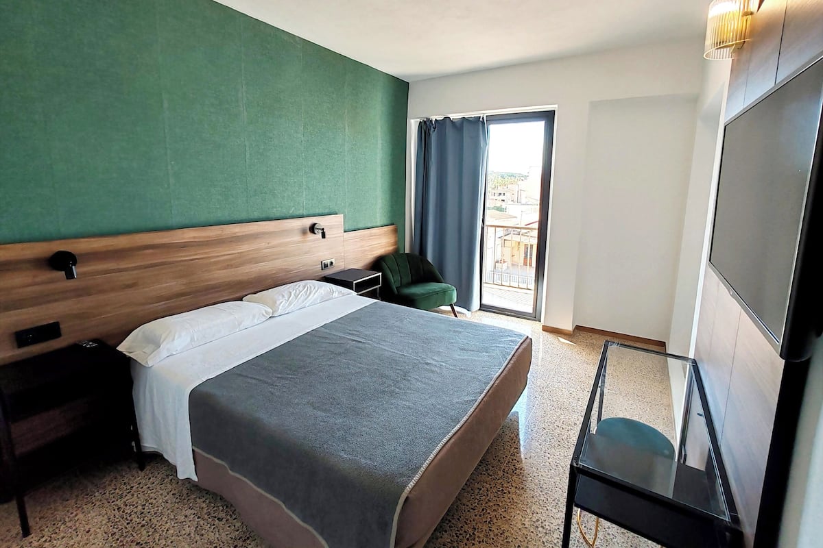 A hotel room with a large bed, a green accent wall, a balcony with a view, and a TV.