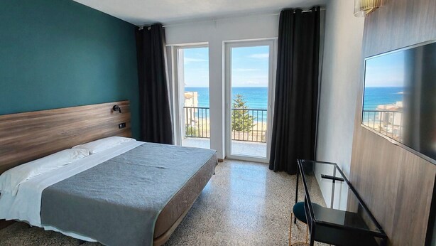 Une chambre d’hôtel équipée d’un lit, d’une télévision, d’un balcon avec vue sur la mer et d’une porte donnant sur l’extérieur.