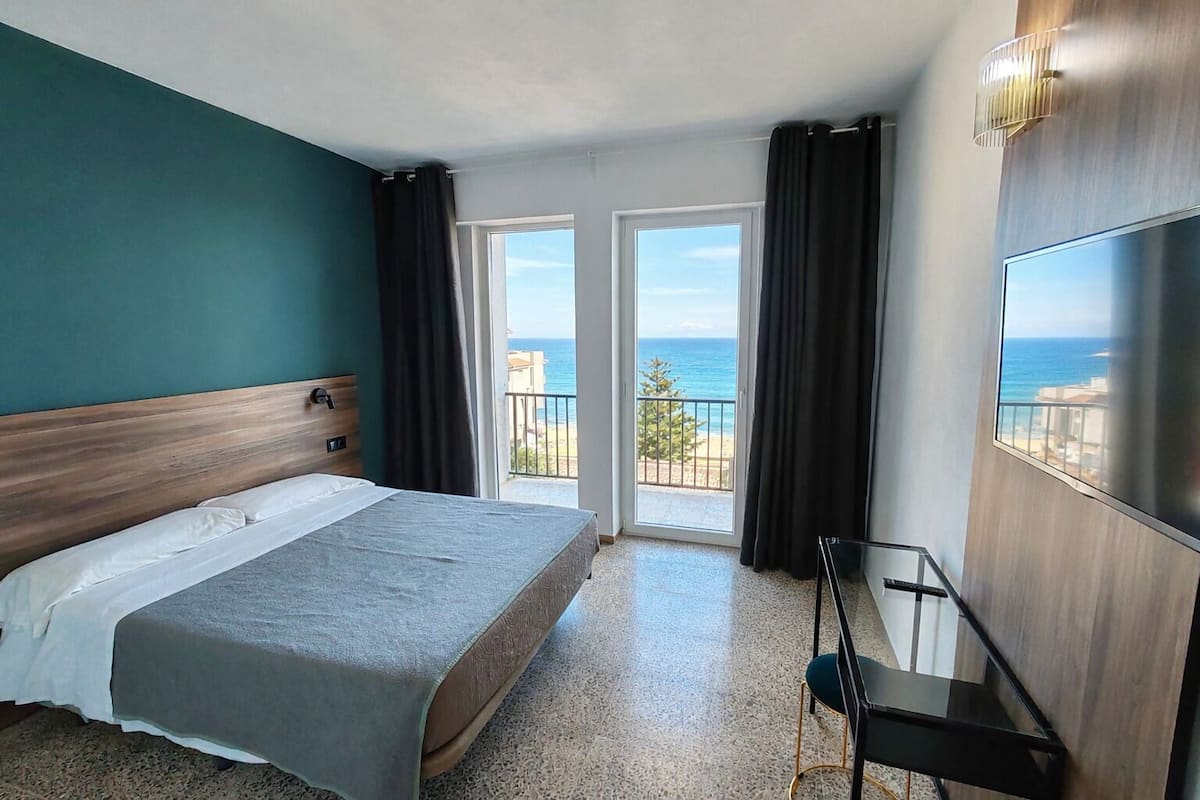 A hotel room with a bed, a TV, a balcony with a view of the sea, and a door leading to the outside.