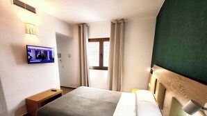 Une chambre d’hôtel avec un lit, un téléviseur fixé au mur, une table de chevet en bois et une fenêtre avec des rideaux.