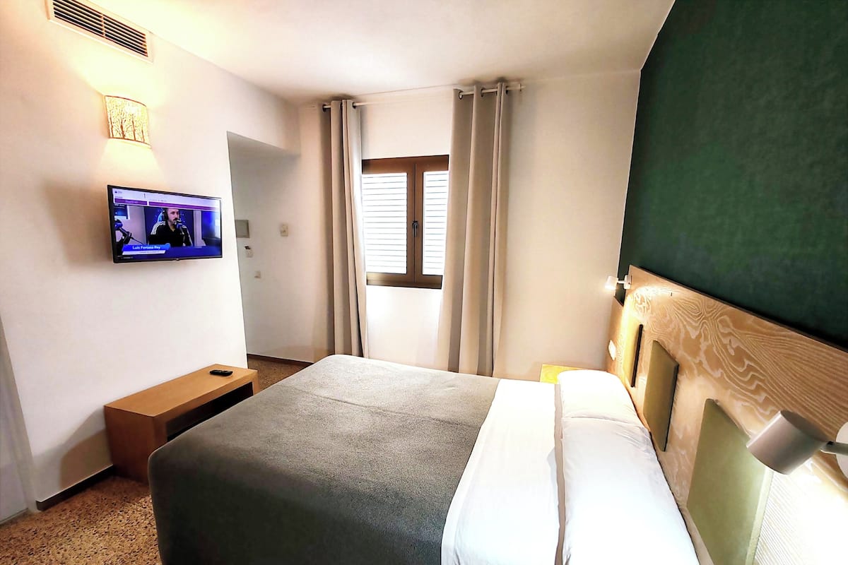 A hotel room with a bed, a TV mounted on the wall, a wooden nightstand, and a window with curtains.