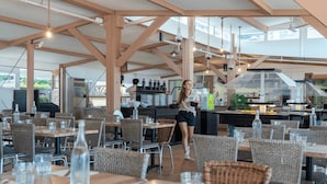 An indoor dining area with wooden tables and wicker chairs, a kitchen in the background, and hanging pendant lights.