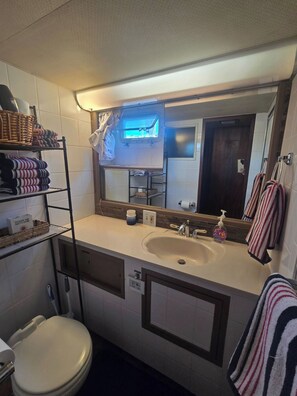 A compact bathroom with a sink, mirror, and toilet. There are shelves with folded towels and a basket on the left. - 3-bedroom house boat in tranquil Key West with AC (Key West)