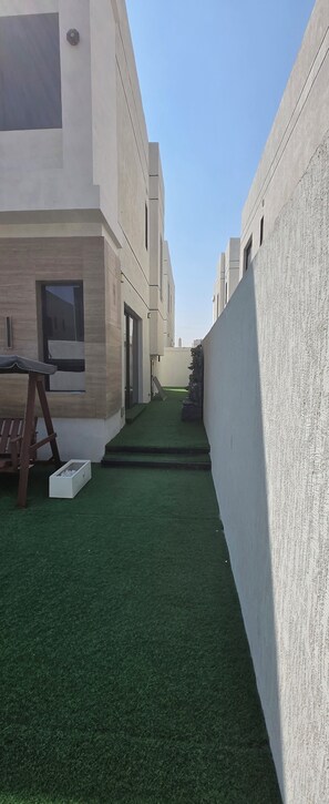 Un edificio moderno con techo plano, un pequeño espacio exterior con césped artificial y un banco de madera.