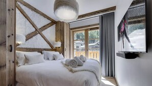 A bedroom with a wooden ceiling, a large bed with white bedding, a television on the wall, and a view of a snowy landscape. - Stunning High-End Apartment with balcons, Les Rives de l’Isère 3 (Val-d'Isère)